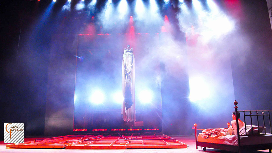 Bright red and white beams of light shine down on Prior Walter lying in bed after the wall has fallen and the Angel decends.