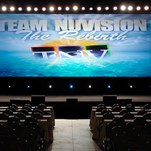 Team Nu Vision Conference with a large rear projection screen up stage with a row of white lights shining downstage for back light.